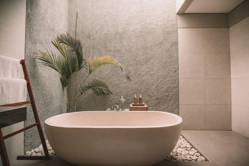 Freestanding soaking tub against a textured greige stone wall with a palm